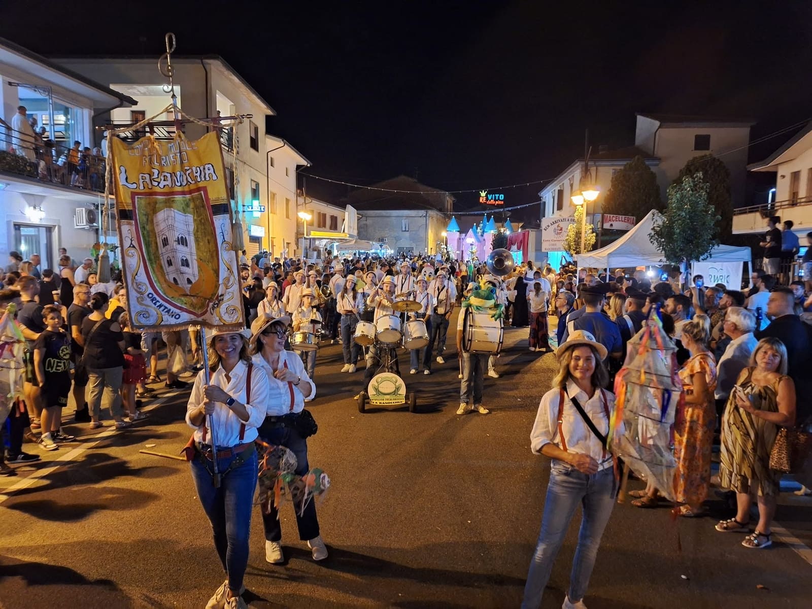 La Ranocchia folk band parading through Orentano on a summer evening, banner raised, drums and frog mascot among the crowd